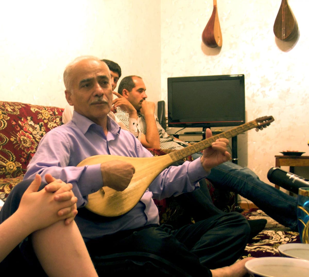 Ismoil Nazriev seated on the floor at home playing a long-necked lute, two younger men behind him.