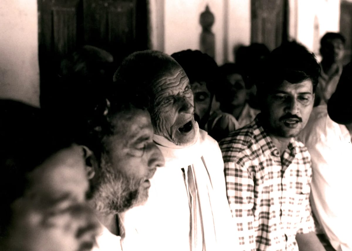 An elderly man singing with his head thrown back among a press of listeners, in a warm-toned photograph.