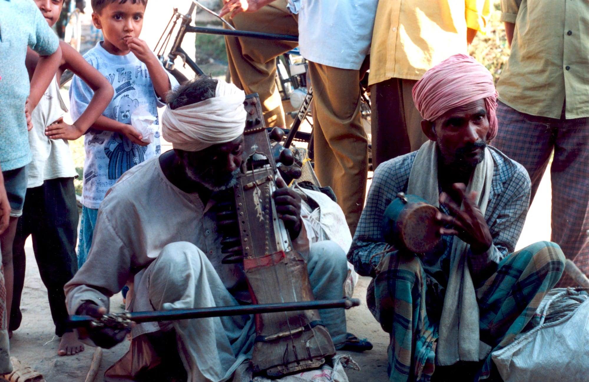 Two street musicians seated on the ground playing a bowed sarangi and small cymbals, a crowd standing around them.
