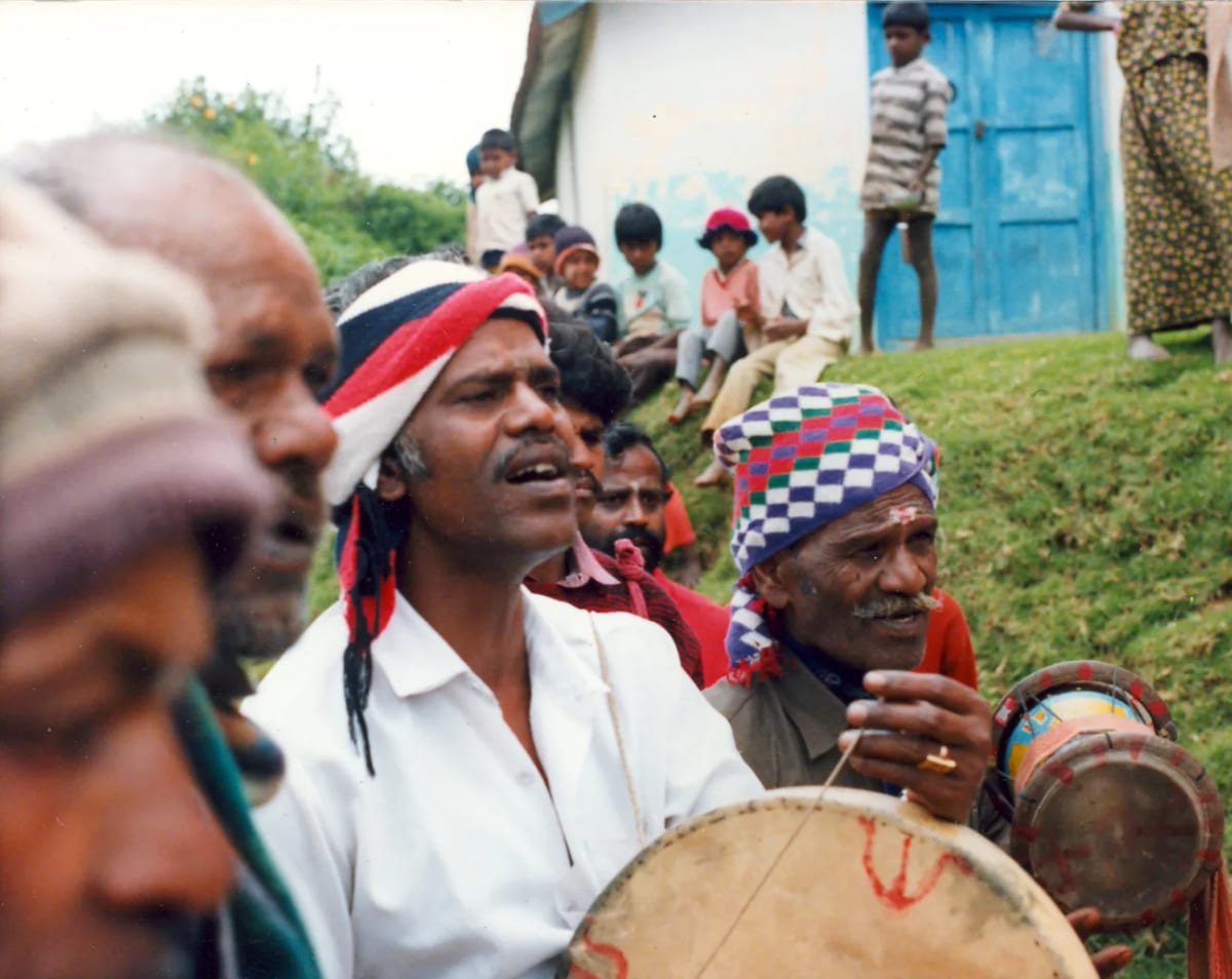 Tamil singers in headcloths performing outdoors with frame drums, villagers and children seated on the slope behind them.