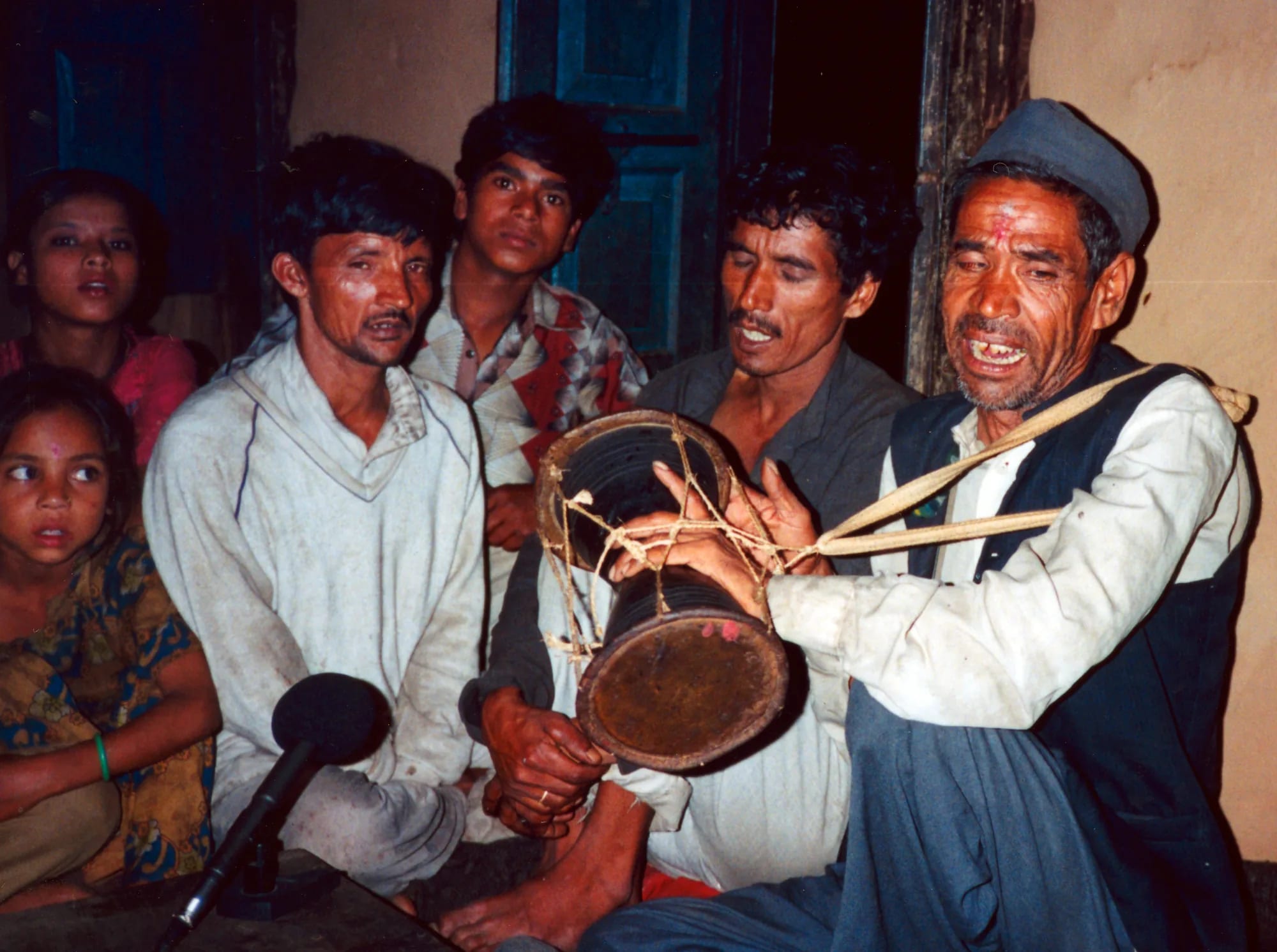 Singers crowded together indoors around a man playing a small hourglass drum, a microphone in the foreground.