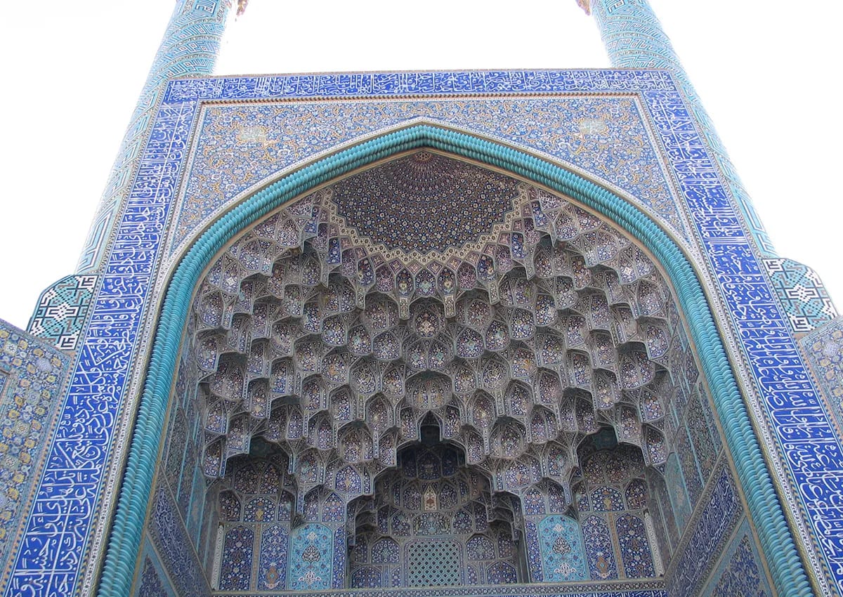 The tiled muqarnas vault of an Iranian mosque portal, seen from below against a pale sky.
