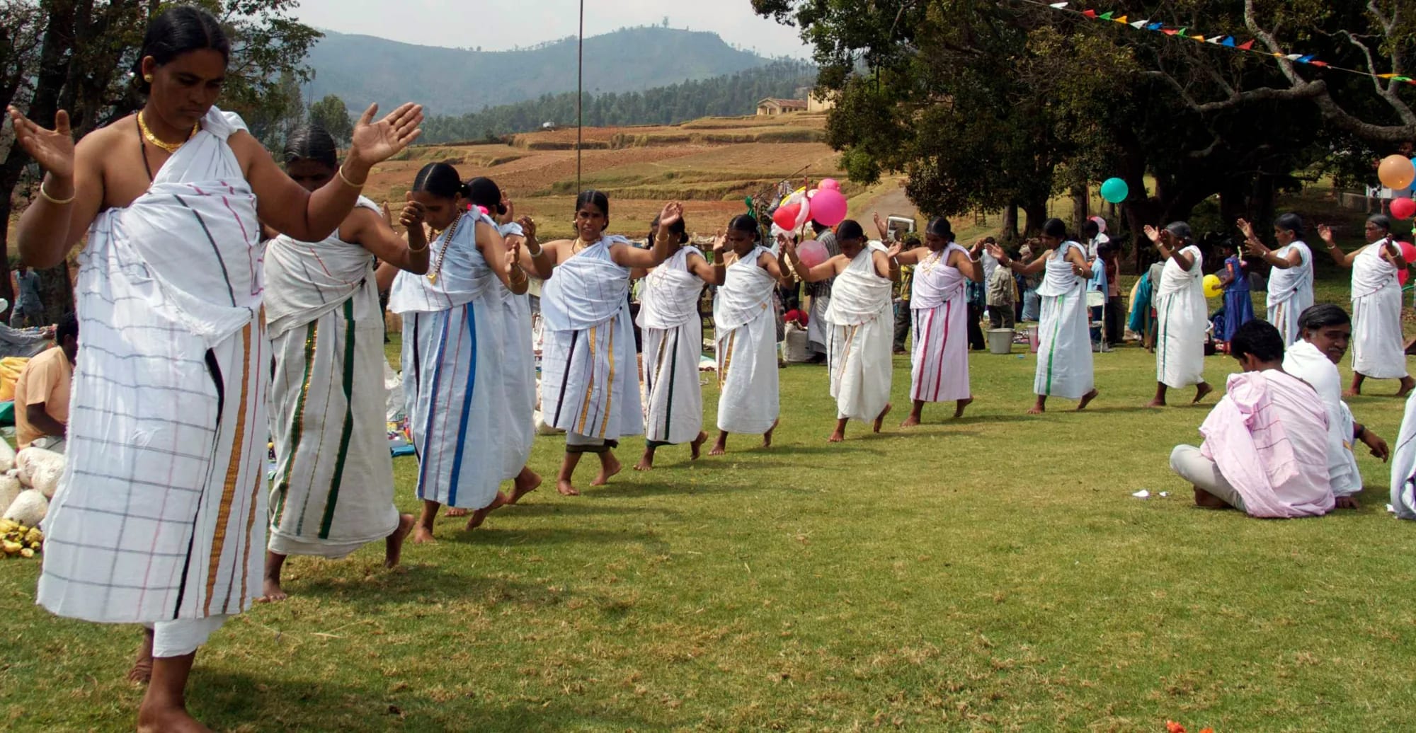 Women in white shawls dancing in a long line on a green hillside meadow, arms raised, watched by a crowd.