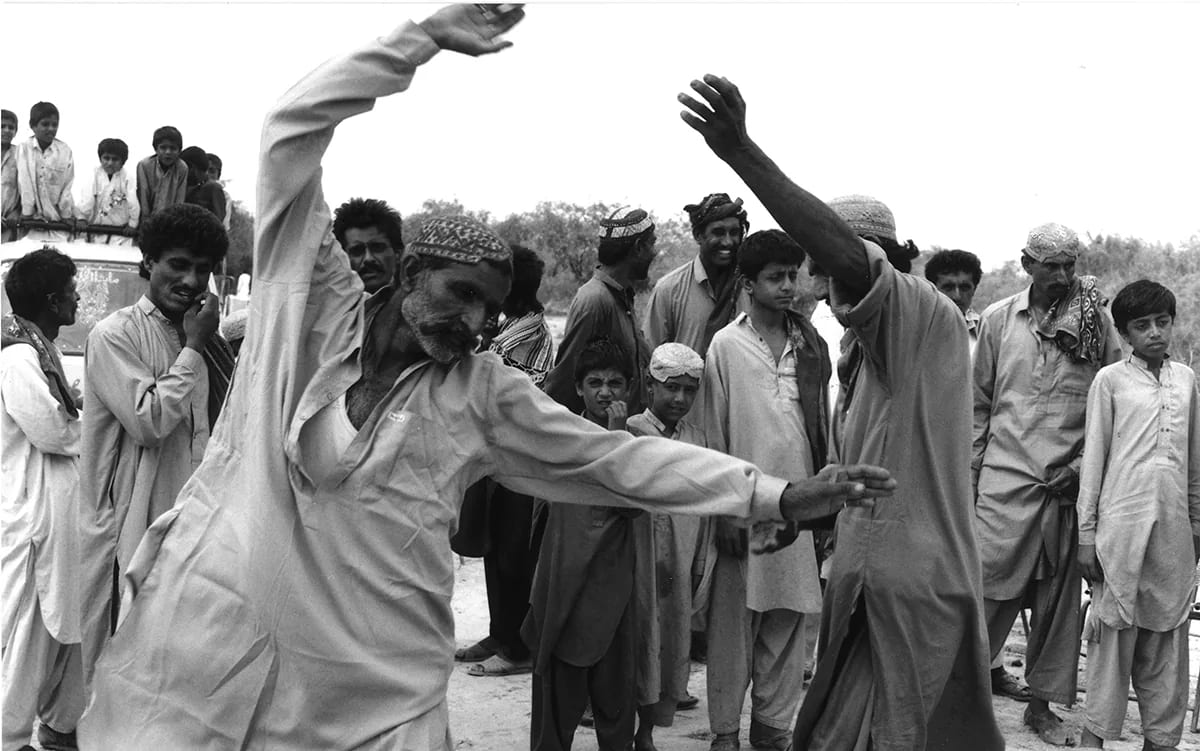 Two men dancing with arms raised, ringed by a crowd of onlookers, in black and white.