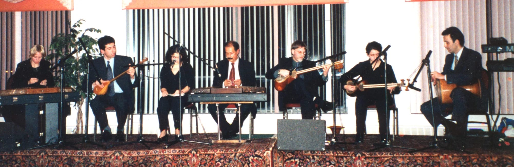 Seven musicians seated in a row on a low stage, playing plucked lutes and a keyboard before a pale curtain.