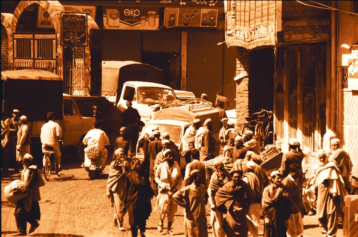 A crowded old-city street of shopfronts, handcarts and parked cars, in warm sepia tones.