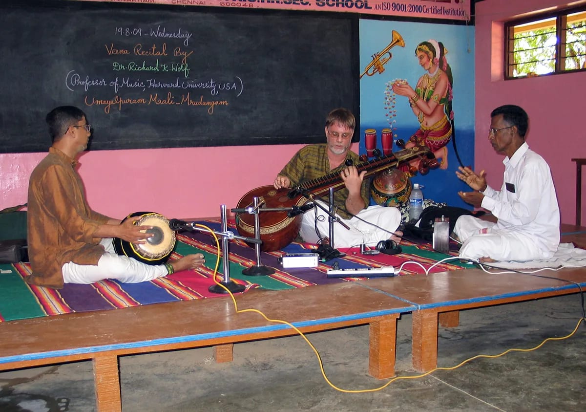 Richard K. Wolf playing the vīṇā on a school stage, flanked by a mridangam player and a singer.