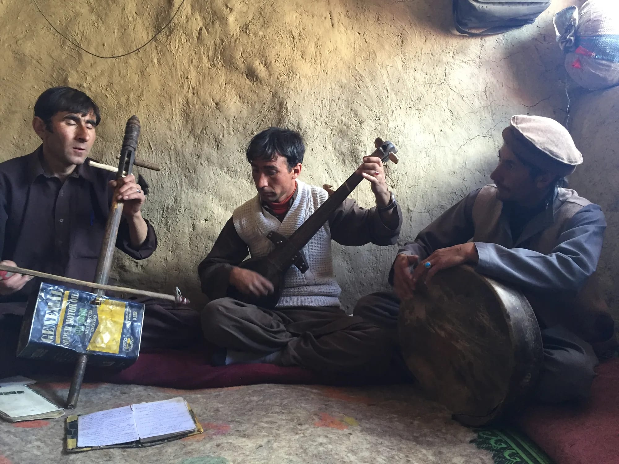 Three Wakhi musicians seated on the floor of a mud-walled room with a bowed lute, a plucked rubab and a frame drum.