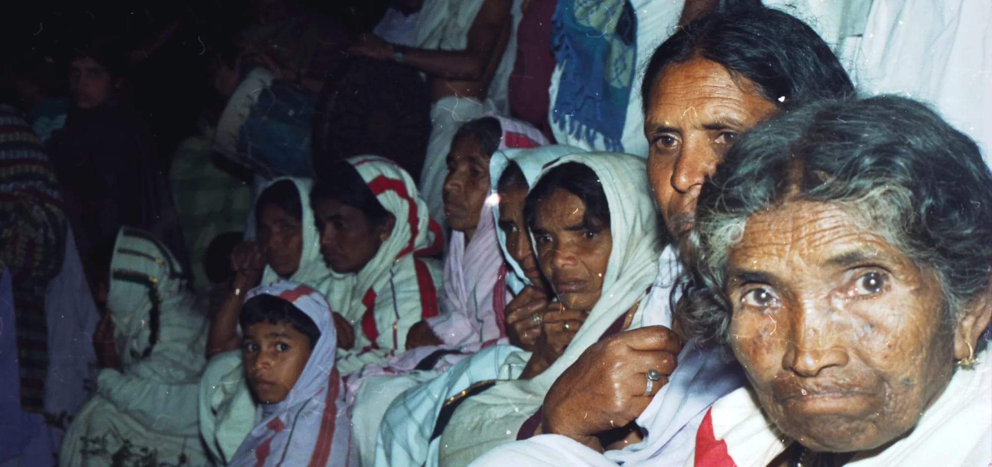 Kota women and children in white shawls seated close together at a funeral, an elderly woman in the foreground.