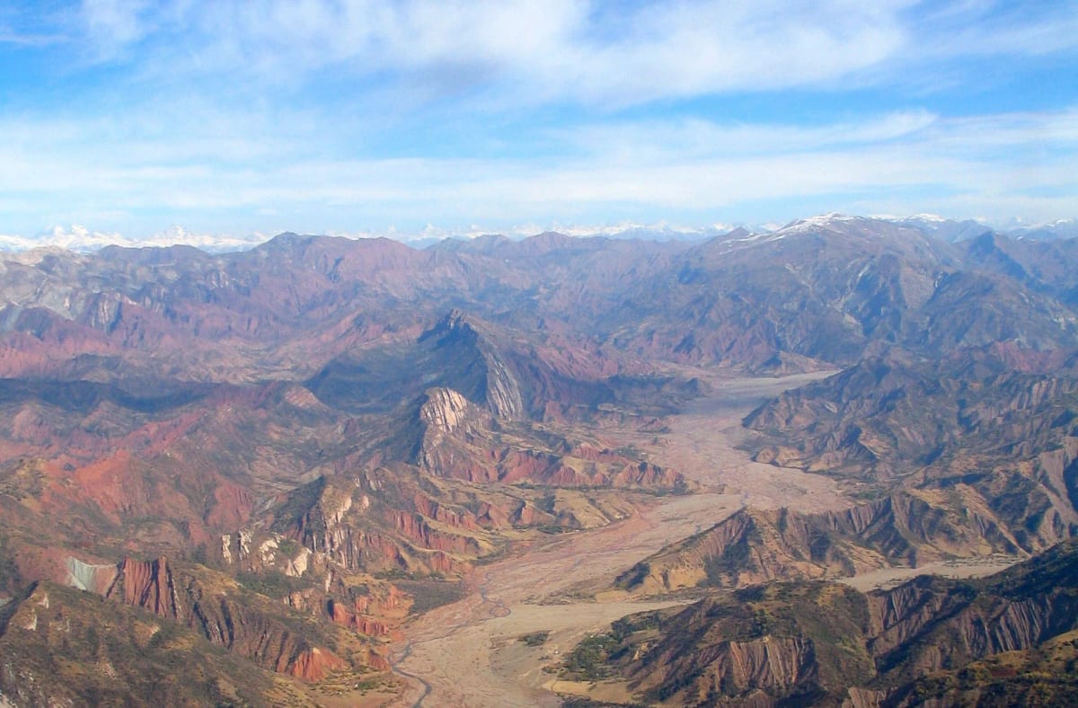 Aerial view of arid red and brown mountain ridges cut by a broad braided riverbed, with snow-capped peaks on the horizon.