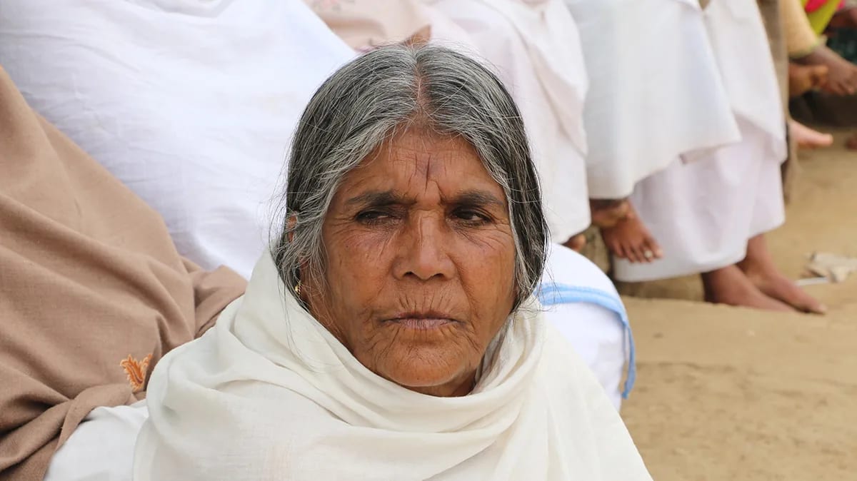 An elderly woman in a white shawl seated on the ground, looking towards the camera.
