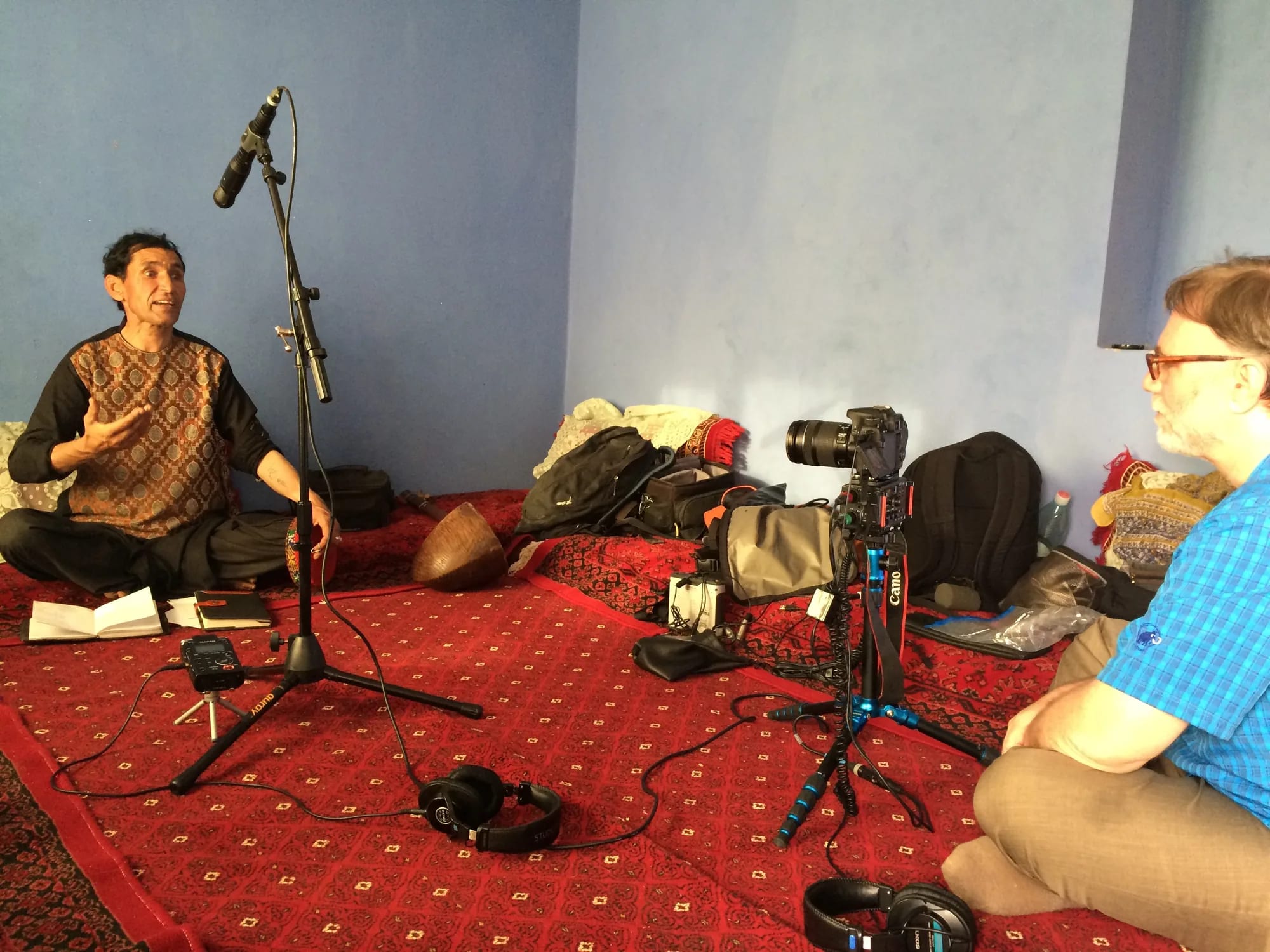 A musician sitting on a red carpet in a blue-walled room, talking across recording microphones and a camera on a tripod to a researcher seated opposite him.