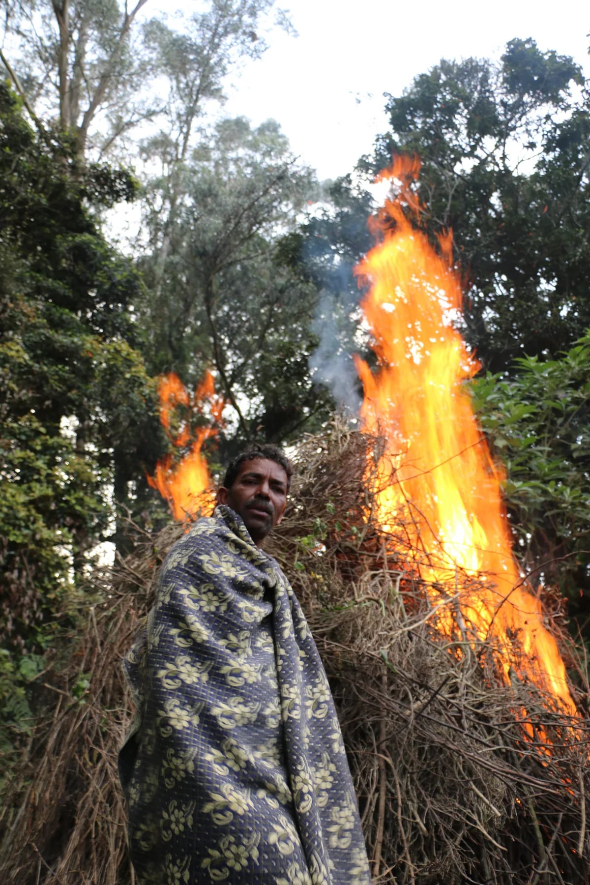 A man wrapped in a patterned blanket standing before a tall bonfire of brushwood at dusk.