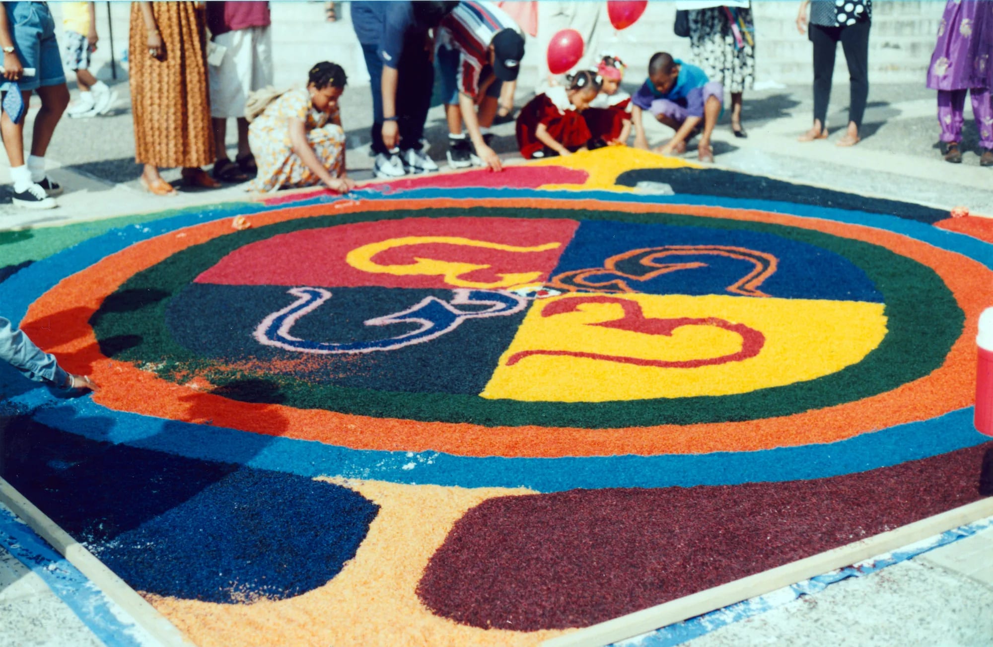 A large circular sand mandala in blocks of colour laid out on a street, with children kneeling at its edge.