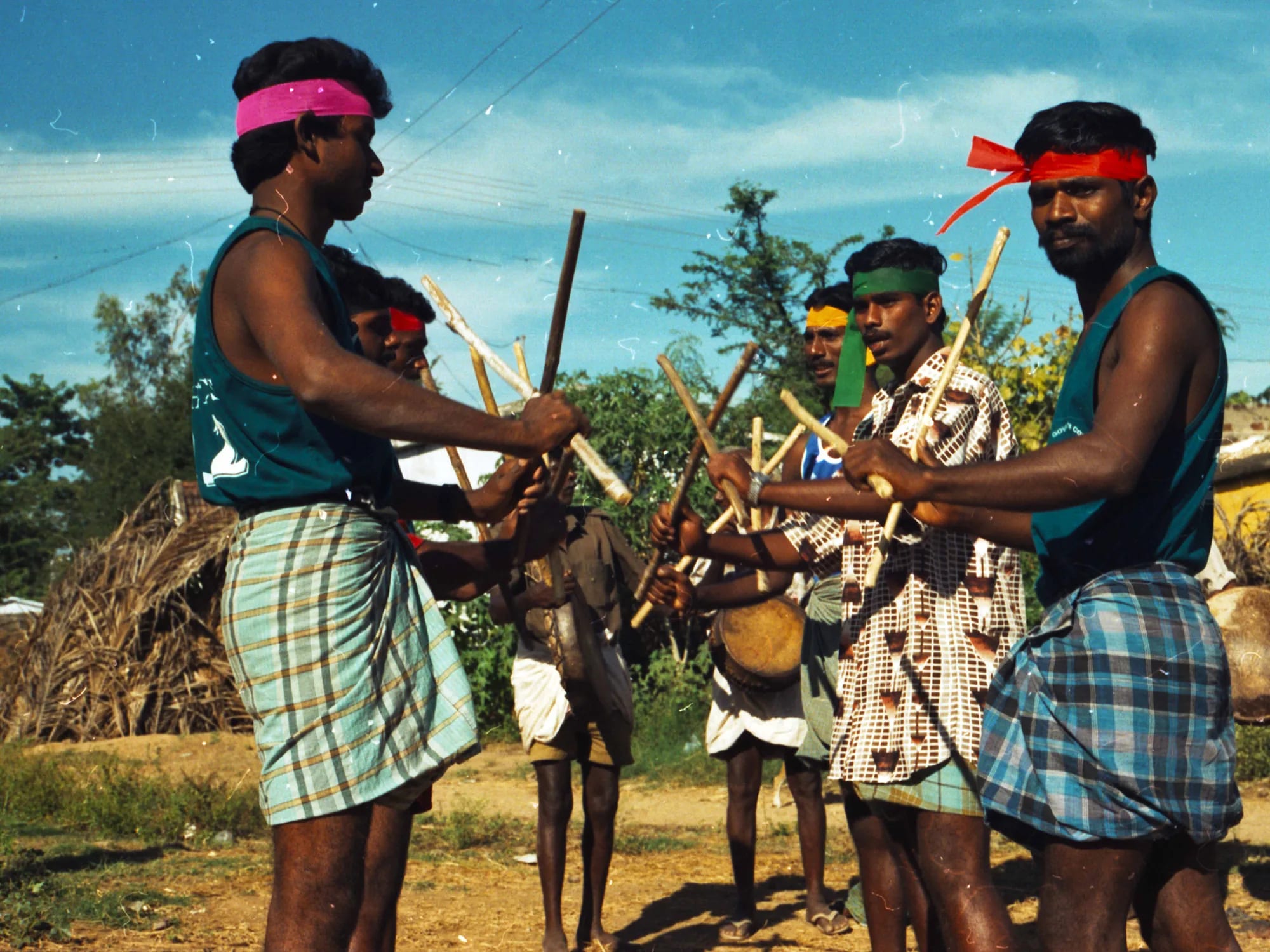 Men in bright headbands striking pairs of wooden sticks together in a village stick dance, a drummer behind them.