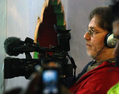 Richard K. Wolf in headphones filming with a shoulder-mounted video camera, a green and red shrine cloth behind him.